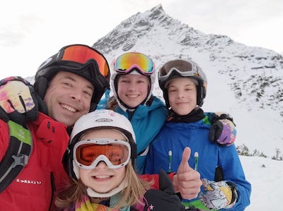 Dad and 3 kids in ski gear with snow covered mountain behind them